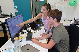Eine Frau zeigt einem Jungen etwas auf einem Computerbildschirm. Beide sitzen an einem Tisch mit Laptops und Büromaterialien. Die Umgebung wirkt modern und hell.
