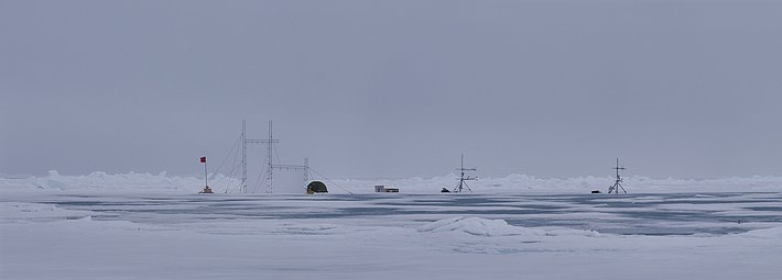Eisige Landschaft mit mehreren Forschungsstationen auf dem Wasser. Sichtbare Strukturen umfassen Masten und Zelte, umgeben von Eis und Wasser.