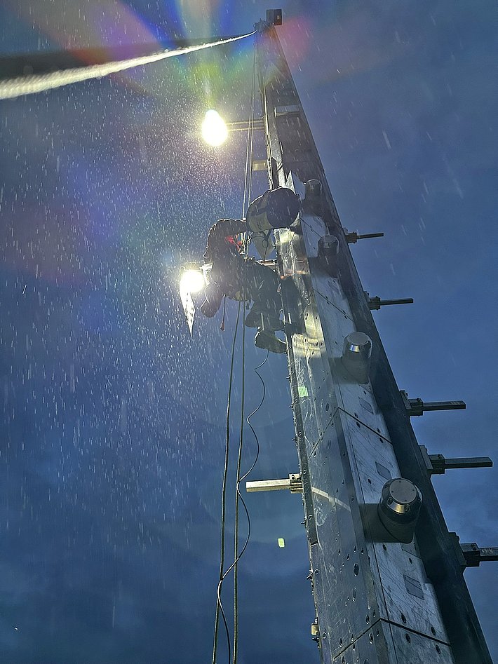 Ein Arbeiter klettert einen hohen Mast hinauf, während es regnet. Der Mast ist mit Scheinwerfern ausgestattet, die Licht in die Szene werfen. Der Himmel wirkt düster und ist von Regen durchzogen.