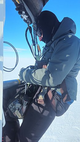 Person in warmer Outdoor-Bekleidung mit Kapuze, Sonnenbrille und Klettergurt, befestigt an Seilen, vor klarem blauen Himmel und Schnee