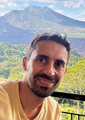 A man with short dark hair smiles at the camera, wearing a light yellow shirt. Behind him, a scenic view features a mountain with a peak and lush greenery in the foreground, indicating a vibrant natural landscape.