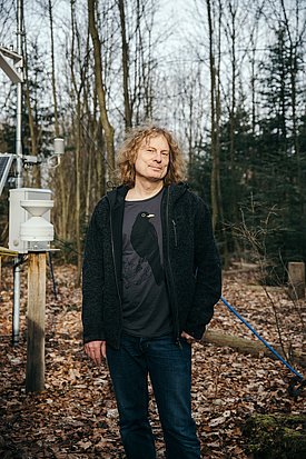 Arthur Gessler steht im Wald, trägt schwarze Jacke, T-Shirt mit Vogelmotiv und Jeans, umgeben von Messgeräten. 