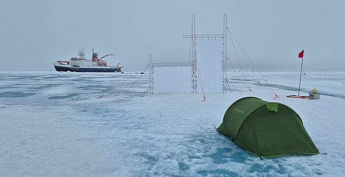 Grünes Zelt auf zugefrorenem See mit roter Flagge, im Hintergrund Forschungsschiff auf Eisfläche.