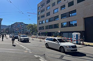 Eine belebte Straße mit gehenden Passanten und Autos, darunter ein weißer Kombi und ein SUV. Symbolbild für Verkehrs- und Baulärm