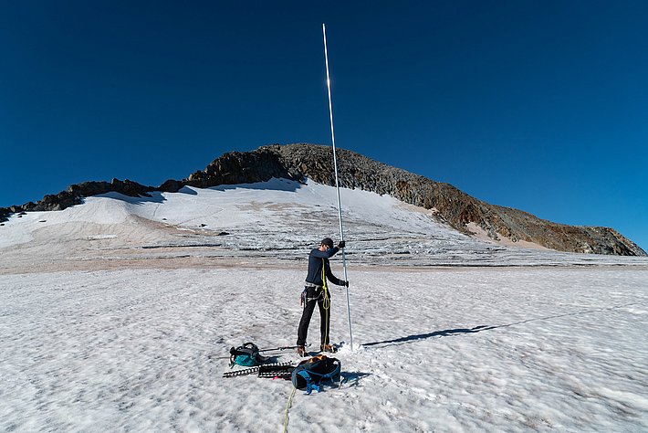Person in outdoor clothing stands on snow-covered glacier holding a long pole, equipment laid on the snow nearby.
