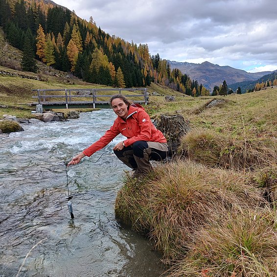 Person in roter Jacke hockt am Ufer eines klaren Bachs in einer herbstlichen Berglandschaft mit Nadelbäumen und Wiesen.