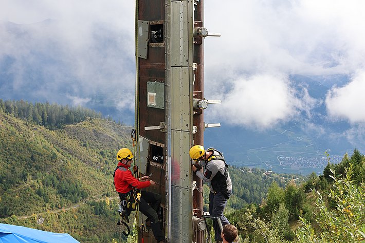 Zwei Personen in Kletterausrüstung besteigen einen hohen, vertikalen Mast in einer bergigen Landschaft. Die Umgebung ist von Bäumen und Wolken umgeben, während die Kletterer die Struktur erklimmen.