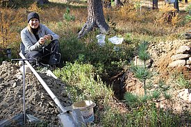Katrin Meusburgn sits on the ground in a forest, next to a freshly planted tree. The surroundings indicate reforestation activity.