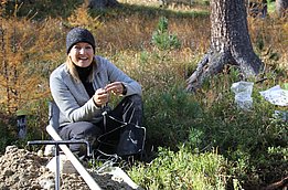 Katrin Meusburgn sits on the ground in a forest, next to a freshly planted tree. The surroundings indicate reforestation activity.