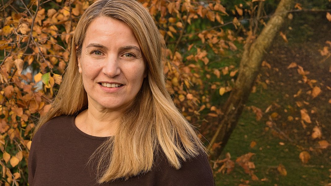 A woman with long brown hair is smiling at the camera, wearing a dark sweater. She stands outdoors surrounded by autumn foliage, with warm tones of orange and yellow in the background. The overall scene conveys a sense of warmth and approachability.