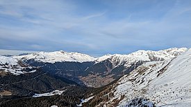 Berglandschaft mit schneebedeckten Gipfeln und bewaldeten Tälern unter bewölktem Himmel.