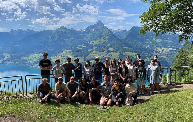 Gruppenfoto von ca. 20 Personen vor Berglandschaft mit See und bewölktem Himmel, aufgenommen an einem Aussichtspunkt mit Geländer