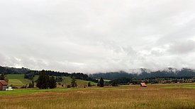 A vast moorland landscape with rolling hills, meadows and trees. The sky is overcast, and thick fog is rolling across the hills in the background. In the foreground lies an autumnal moor.