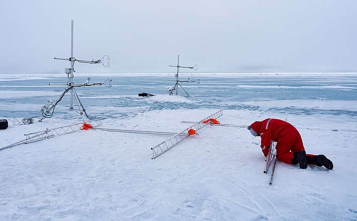 Zwei Windmessstationen auf verschneitem Boden, verbunden mit einem Gerüst, daneben eine Person in rotem Anzug, die am Gerüst arbeitet.
