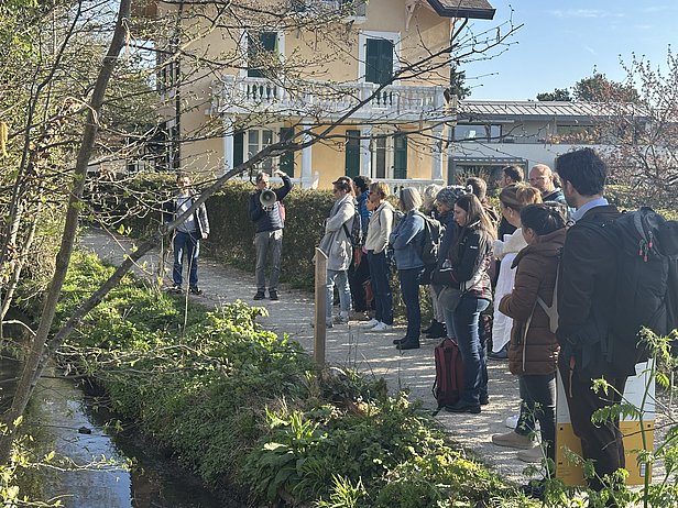 Gruppe von Menschen hört einer Person zu, die neben einem kleinen Bach vor einem gelben Haus spricht, Frühlingsbäume im Hintergrund.