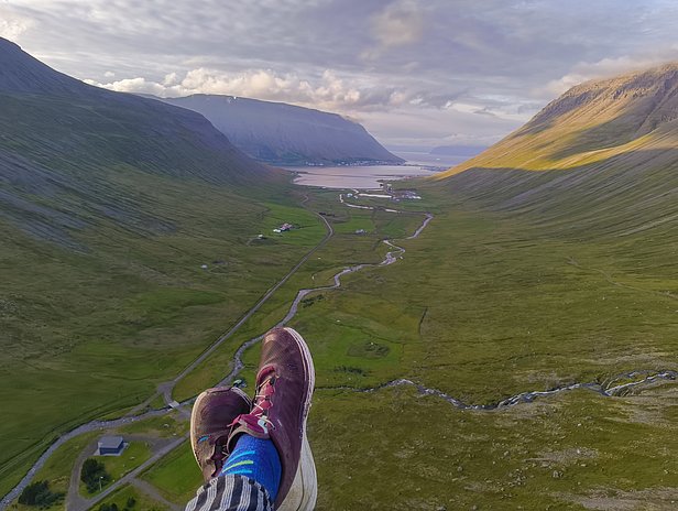 Die Aufnahme zeigt einen Blick von einer hohen Stelle auf ein weites Tal mit sanften Hügeln und einem Fluss, der durch das Grün fließt. Im Vordergrund sind ein Paar Beine mit bunten Socken und Sportschuhen zu sehen. Die Landschaft strahlt Ruhe und Weite aus.