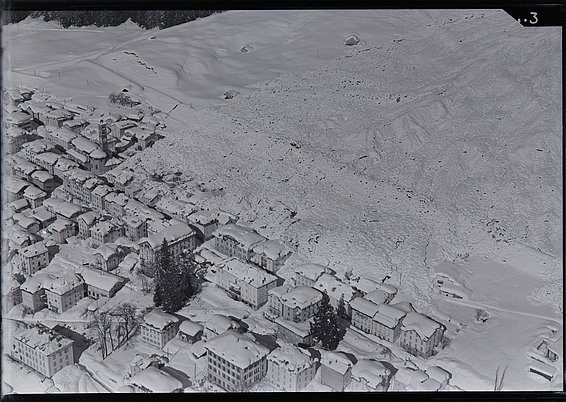 Vue aérienne en noir et blanc d'un cône d'avalanche sur un versant montagneux à côté d'un village avec des bâtiments et des routes