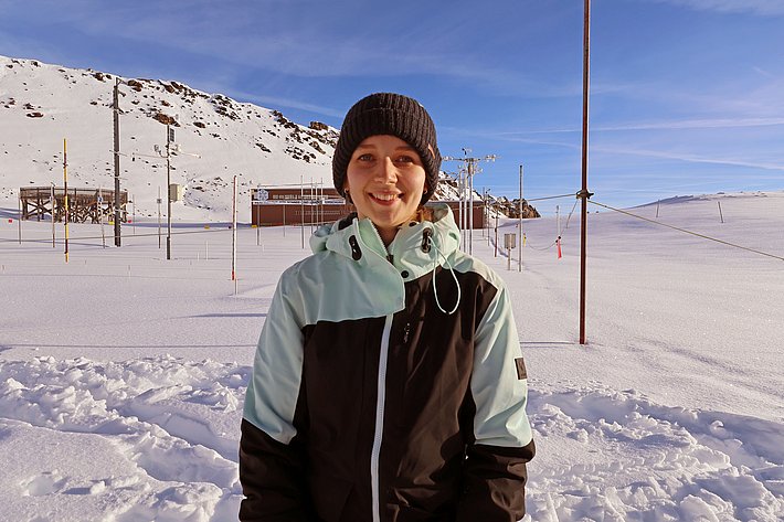 Eine junge Person steht in einem schneebedeckten Landschaftsbereich. Sie trägt eine Winterjacke und eine Mütze und lächelt freundlich. Im Hintergrund sind Forschungsanlagen und Berge zu erkennen, die den winterlichen Kontext verdeutlichen.