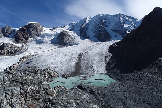 Das Bild zeigt eine majestätische Berglandschaft mit Gletschern, Fels und einem grünen Gletschersee. Der Himmel ist blau und die Sonne scheint.  