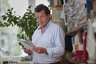 Paolo Cherubini von der WSL steht in einem Raum, hält Papiere in der Hand und schaut direkt in die Kamera. Im Hintergrund sind Regale mit Dokumenten und eine Pflanze sichtbar. Die Atmosphäre wirkt geschäftig und kreativ.