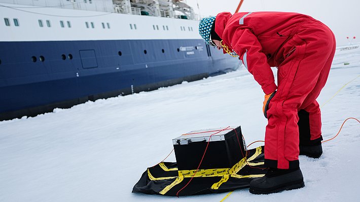Person in rotem Overall und blauer Mütze beugt sich über schwarze Kiste auf Schlitten vor einem Schiff im Schnee.