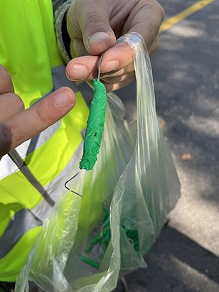Mano che tiene una larva verde di plastilina su un filo di ferro, con una busta di plastica trasparente e un indumento riflettente sullo sfondo.