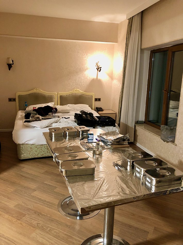 The image shows an unmade bed with scattered clothes and personal items next to a dining table covered with aluminium foil. Several metal containers are placed on the table in a hotel room with warm lighting and a window nearby.