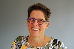 A woman with short hair and glasses is smiling into the camera. The background is grey and neutral.