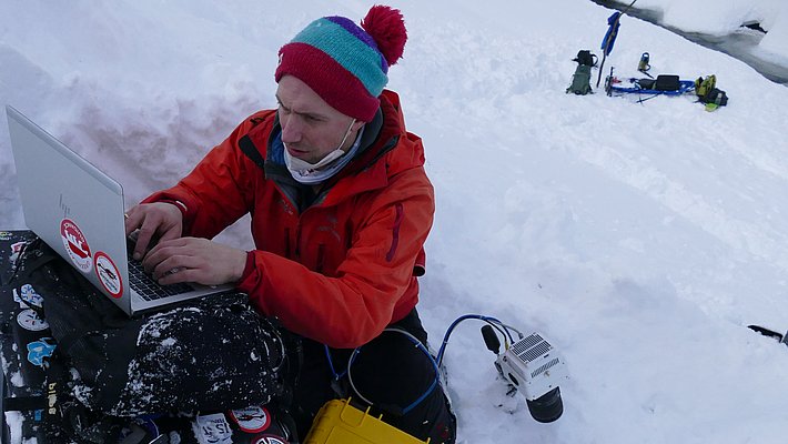 Un homme assis sur la neige utilise un ordinateur portable, vêtu d'une veste rouge et portant un bonnet coloré. Il travaille devant une boîte noire couverte d'autocollants. L'environnement est enneigé, avec des équipements visible au fond.