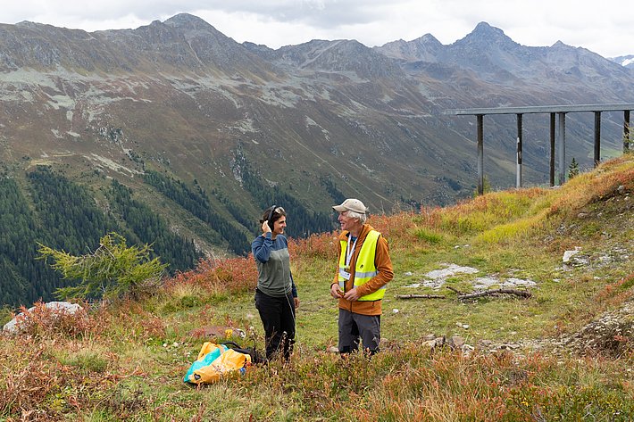 Zwei Personen stehen auf einer Wiese mit buntem Gras und Blick auf die Berge. Eine Person trägt eine Weste und hält ein Notizbuch, die andere hat Kopfhörer auf.