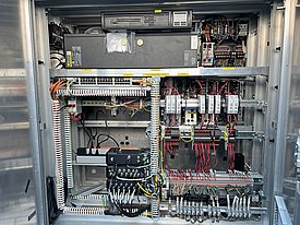 Blick in ein metallisches Gehäuse mit Kabeln, Anschlüssen und Schaltgeräten. Die innere Komponente enthält eine Vielzahl von Leitungen und Modulen, die zur Steuerung von elektrischen Systemen verwendet werden.