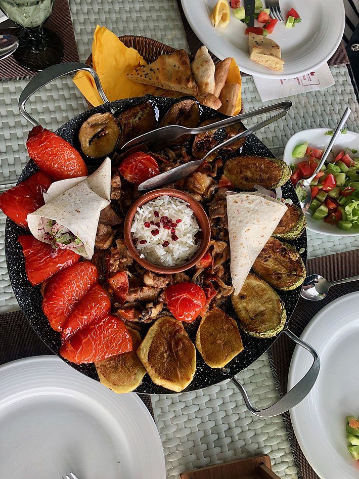 A large, round platter showcases a variety of grilled meats, vibrant roasted vegetables, and flatbreads. A small bowl of white sauce sits in the center, surrounded by garnished tomatoes and slices of red and yellow ingredients, creating a colorful and appetizing presentation.