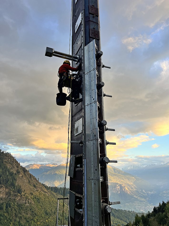 Ein Arbeiter in Sicherheitsausrüstung klettert an einer hohen, stählernen Struktur entlang. Im Hintergrund sieht man eine eindrucksvolle Landschaft mit Bergen und einem bewölkten Himmel.
