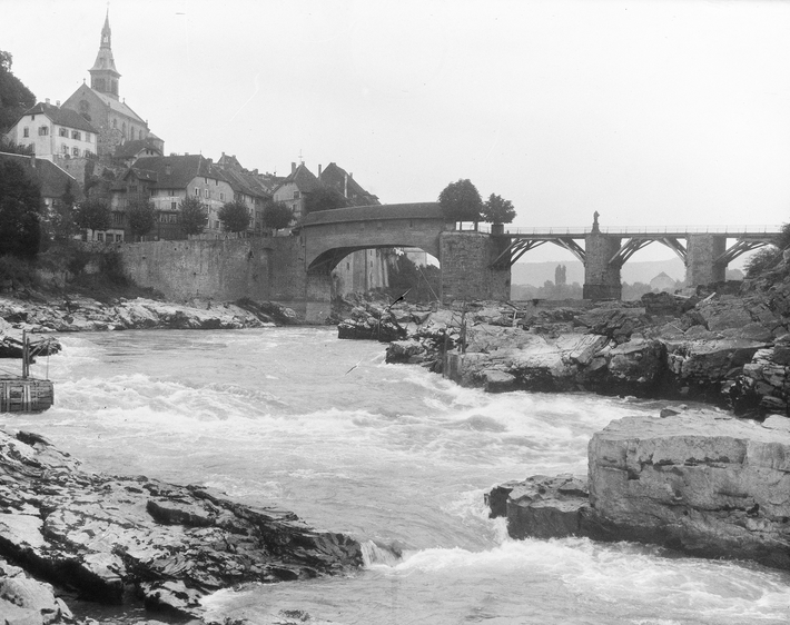 The Rhine with rocks and fast-flowing water is in the foreground, with a bridge and Laufenburg visible in the background.