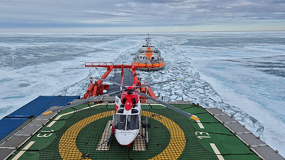 Hubschrauber auf grünem Landeplatz eines Schiffes, das durch eine Eislandschaft fährt, mit einem weiteren Schiff im Hintergrund.