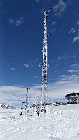 Hoher weißer Messmast mit Wetterstation auf schneebedecktem Gelände unter blauem Himmel, im Hintergrund ein Gebäude auf Stelzen