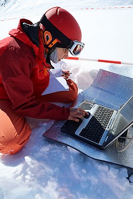 Anja Mödl in rotem Skihelm und rotem Skianzug arbeitet mit Laptop und Notizblock im Schnee