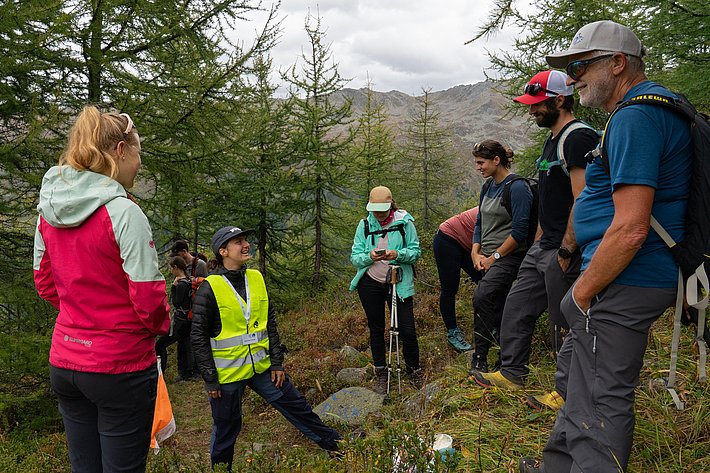 Personengruppe in der Natur, umgeben von Nadelbäumen. Eine Person trägt eine gelbe Warnweste, eine andere hält ein Smartphone. Die anderen sind in Freizeitkleidung.