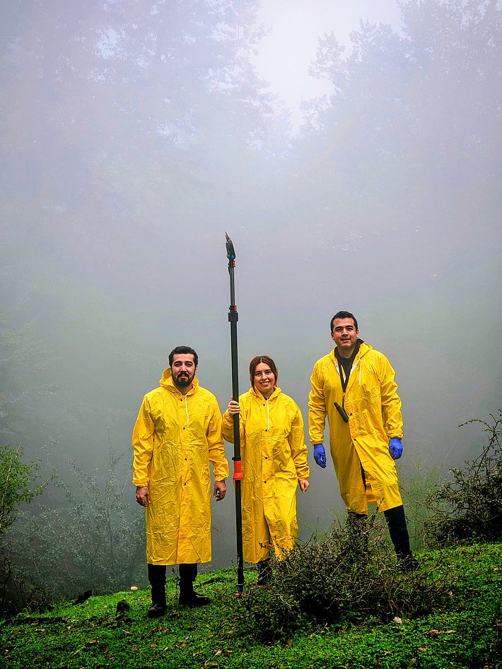 Three individuals stand on a grassy area, wearing bright yellow raincoats and gloves. They are positioned in a foggy environment, with trees faintly visible in the background. One person holds a long tool, highlighting their readiness for outdoor work or an environmental task.