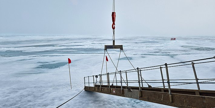 Metallbrücke mit Geländer führt über zugefrorenen See, darüber hängt ein Kranhaken, rote Fahnen markieren Stellen im Eis.