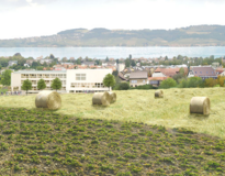 Ein fiktionaler Ort im Seeland heute. Man sieht Häuser, im Vordergrund Ackerland und Wiese mit Heuballen. Im Hintergrund der See und Hügel.