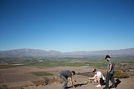 Drei Männer begutachten die Bodenverhältnisse auf einem trockenen Feld mit Blick auf ein Tal und Berge im Hintergrund