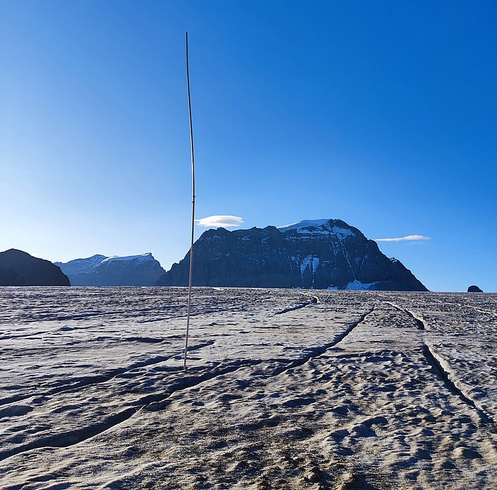 Snow-covered terrain with a thin, tall pole in the center and a mountain massif in the background under a clear sky.