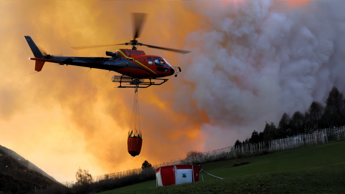 Hubschrauber mit rotem Löschbehälter fliegt vor orangefarbenem Rauch über grünem Feld mit Feuerwehrzelt.