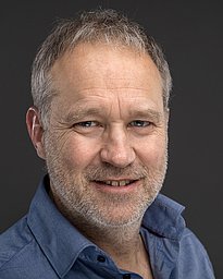 A middle-aged man with short, graying hair and a stubbled beard smiles at the camera. He is wearing a blue button-up shirt, and the background is a neutral gray, highlighting his friendly expression.