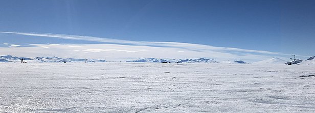 Eine weite, schneebedeckte Landschaft unter blauem Himmel. Im Hintergrund sind schneebedeckte Berge zu sehen. Der gesamte Bereich wirkt kalt und unberührt, mit wenigen Spuren menschlicher Aktivität.