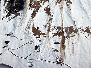 Schneebedeckter Berg mit sichtbaren Gleitschneelawinen-Spuren und kleinen Gebäuden am Fuß des Hangs unter klarem blauem Himmel