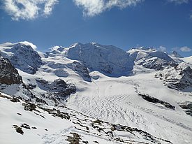 A panoramic view of majestic snow-covered mountains under a clear blue sky. The landscape features glaciers and rugged terrain, showcasing the beauty of a winter alpine environment. Snow blankets the ground, creating a serene and untouched atmosphere.