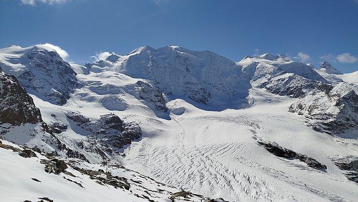 A panoramic view of majestic snow-covered mountains under a clear blue sky. The landscape features glaciers and rugged terrain, showcasing the beauty of a winter alpine environment. Snow blankets the ground, creating a serene and untouched atmosphere.