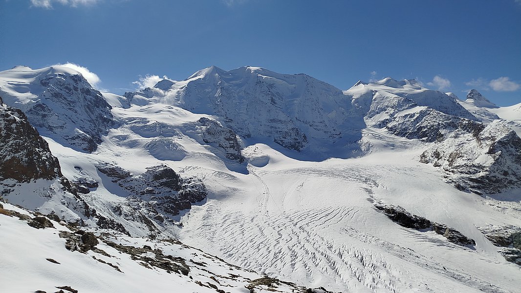 A panoramic view of majestic snow-covered mountains under a clear blue sky. The landscape features glaciers and rugged terrain, showcasing the beauty of a winter alpine environment. Snow blankets the ground, creating a serene and untouched atmosphere.